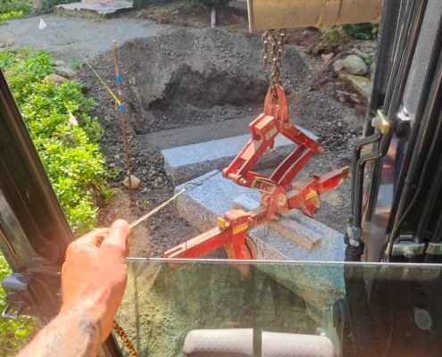 View from the cab of a mini excavator picking up granite steps with a red multi lifting device.