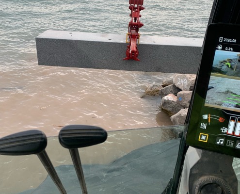 Mini excavator sets a long granite monument into the water for shoreline erosion control.