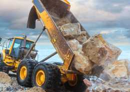 A large yellow dump truck poured heavy boulders onto the ground with the ocean and grey sky in the background.