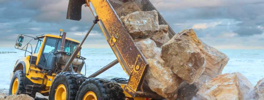 A large yellow dump truck poured heavy boulders onto the ground with the ocean and grey sky in the background.