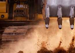 The metal bucket teeth of a backhoe as it picks up mounds of dirt with the operating cab visible in the background.