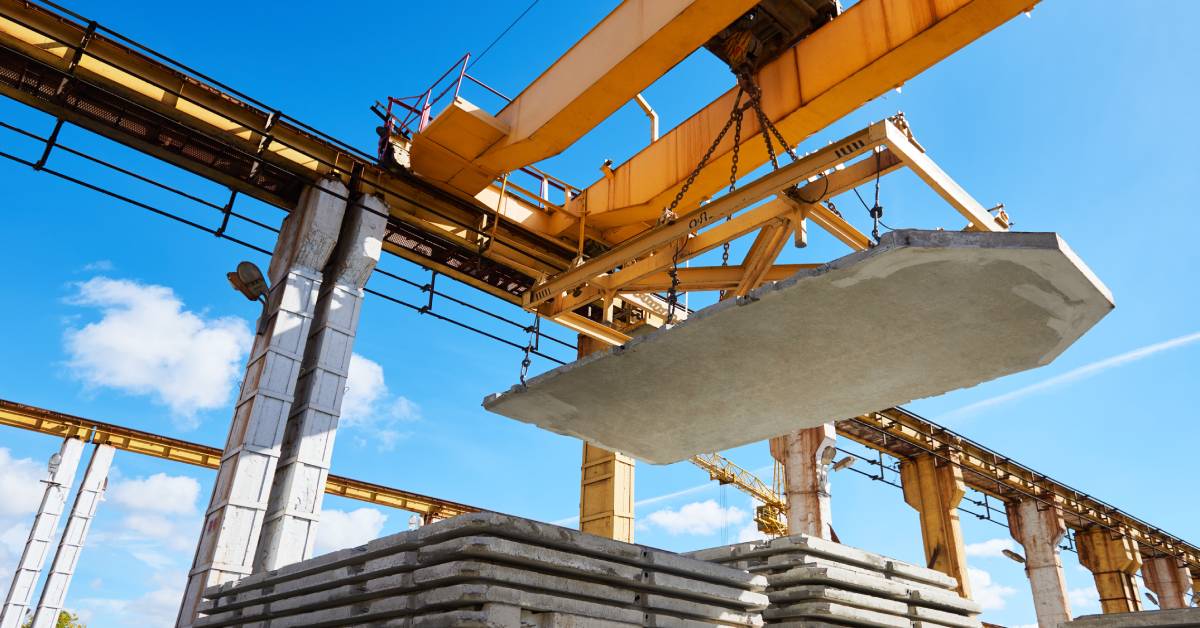 An underneath view of a yellow industrial slab lifter hoisting up a large concrete slab from a stack of other concrete slabs.