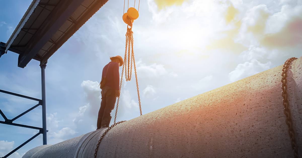 A worker in a hard hat and protective mask standing on top of a large concrete pipe with chains wrapped around it.