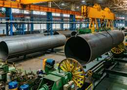 A factory warehouse with multiple, massive steel pipes and one of them being lifted in the air by hooks.