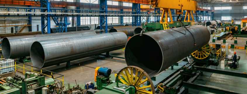 A factory warehouse with multiple, massive steel pipes and one of them being lifted in the air by hooks.