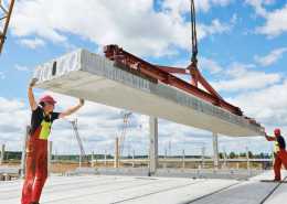 Two construction workers in red gear standing on top of an unfished building using industrial machinery to move a concrete slab.
