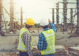 Two male architects in high-visibility vests and hard hats are pointing at an unfinished construction site.