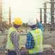 Two male architects in high-visibility vests and hard hats are pointing at an unfinished construction site.