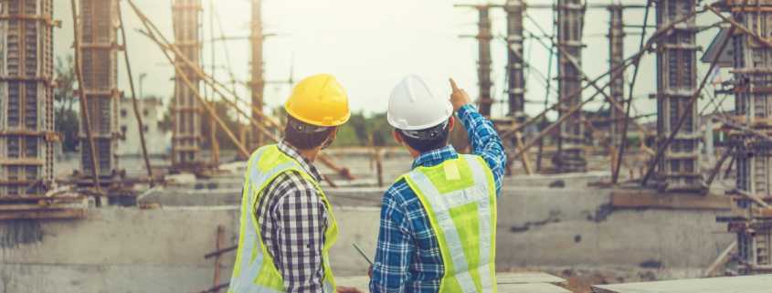 Two male architects in high-visibility vests and hard hats are pointing at an unfinished construction site.