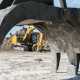 A close-up of a large, grey excavator attachment covered in sand on a construction site with other excavators in the background.