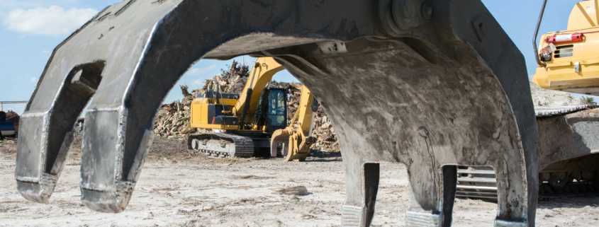 A close-up of a large, grey excavator attachment covered in sand on a construction site with other excavators in the background.