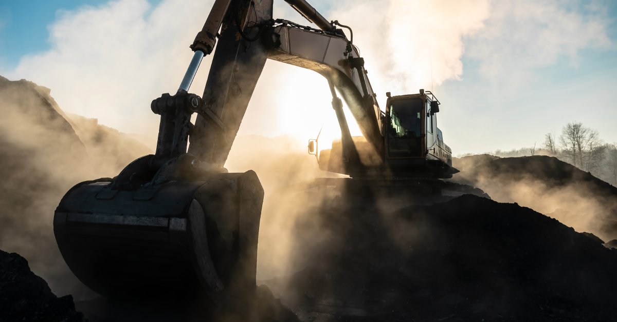 A shadowy, cinematic view of a yellow excavator and bucket attachment on top of mounds of dirt under a bright blue sky.