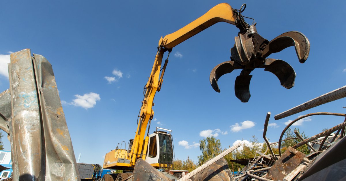 An underneath view of a yellow excavator with a grapple attachment to pick up pieces of scrap metal.