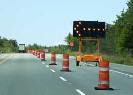 A backroad highway cutting through dense forestry with a line of barrel cones in the middle and a light arrow pointing left.