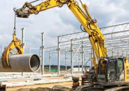 A yellow excavator with a pipe lifter attachment lifting up a large concrete pipe from a mound of dirt.