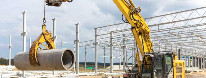 A yellow excavator with a pipe lifter attachment lifting up a large concrete pipe from a mound of dirt.