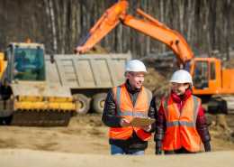 A male and female worker in high visibility gear in front of construction equipment. The man is smiling and holding a clipboard.