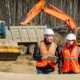 A male and female worker in high visibility gear in front of construction equipment. The man is smiling and holding a clipboard.