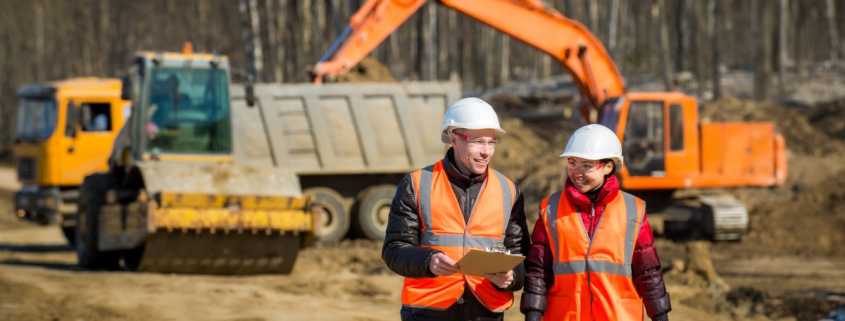 A male and female worker in high visibility gear in front of construction equipment. The man is smiling and holding a clipboard.