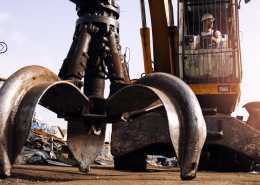 A man in a white hard hat inside the operator's cabin of an excavator. The excavator has a claw attachment.