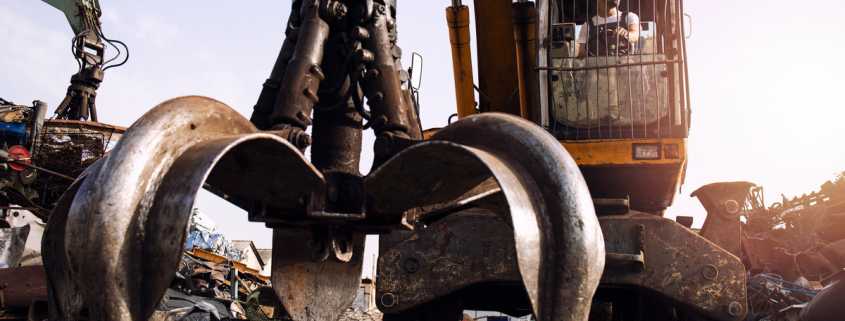 A man in a white hard hat inside the operator's cabin of an excavator. The excavator has a claw attachment.