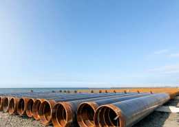 A row of long steel pipes sitting on a bright, sunny seashore. The pipes are on short concrete stands.