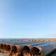 A row of long steel pipes sitting on a bright, sunny seashore. The pipes are on short concrete stands.