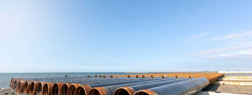 A row of long steel pipes sitting on a bright, sunny seashore. The pipes are on short concrete stands.