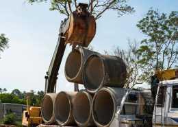 An excavator using a pipe hook to lift concrete pipes from a stack of pipes piled on the back of a large truck.