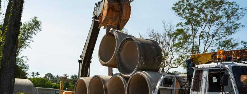 An excavator using a pipe hook to lift concrete pipes from a stack of pipes piled on the back of a large truck.