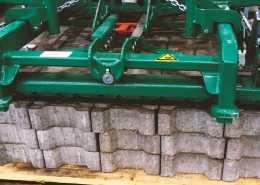 Concrete bricks on a wooden pallet about to be lifted by a green, mechanical lifting machine.
