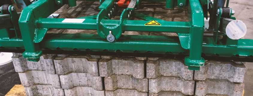 Concrete bricks on a wooden pallet about to be lifted by a green, mechanical lifting machine.