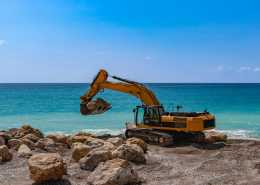 A yellow excavator on a beach near the ocean uses its bucket to pick up one of several boulders in the area.
