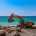 A yellow excavator on a beach near the ocean uses its bucket to pick up one of several boulders in the area.