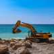 A yellow excavator on a beach near the ocean uses its bucket to pick up one of several boulders in the area.