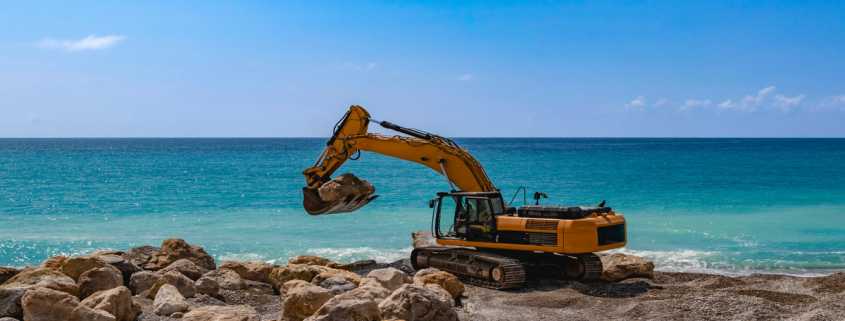 A yellow excavator on a beach near the ocean uses its bucket to pick up one of several boulders in the area.