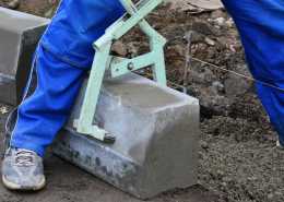 An individual wearing blue pants and sneakers while using a green lifting tool to lift a large block of concrete.