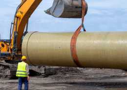 A construction worker standing near an excavator bucket that is lifting a large pipe on a muddy construction site.