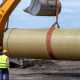 A construction worker standing near an excavator bucket that is lifting a large pipe on a muddy construction site.