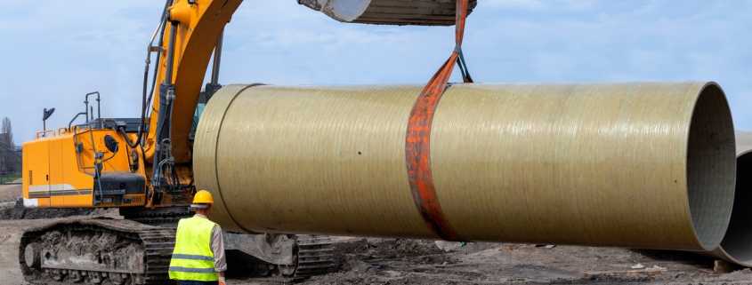 A construction worker standing near an excavator bucket that is lifting a large pipe on a muddy construction site.