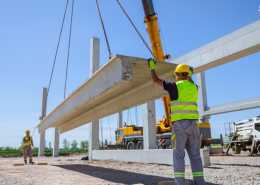 Two construction workers in safety vests and hard hats guiding a crane lifting a large slab of concrete.