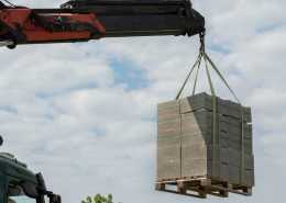 A hydraulic crane using a hook and green ropes to lift several stacks of concrete blocks in the air.