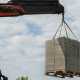 A hydraulic crane using a hook and green ropes to lift several stacks of concrete blocks in the air.