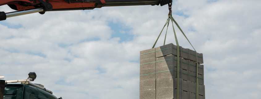 A hydraulic crane using a hook and green ropes to lift several stacks of concrete blocks in the air.