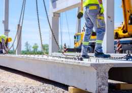 Two construction workers attaching chains to a large piece of concrete with heavy equipment in the background.