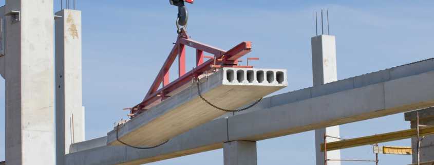 A large slab of concrete is being lifted in the air by lifting attachments. There are concrete columns in the background.