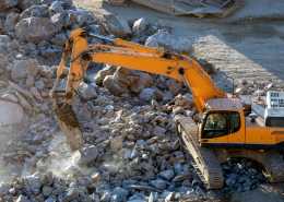 A large yellow heavy machine is on a construction site on top of rocks, using its arm to lift the large rocks.