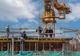 A construction site has heavy machinery surrounded by workers in hard hats. There is a lifting attachment hanging in the air.
