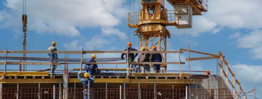 A construction site has heavy machinery surrounded by workers in hard hats. There is a lifting attachment hanging in the air.