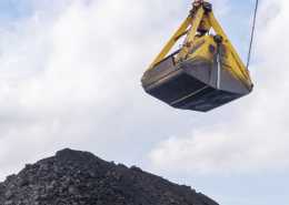 A large yellow excavator crane bucket hovers above a pile of black coal under a cloudy sky at a mining or construction site.